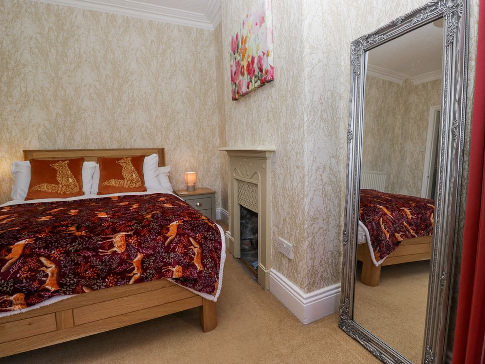 A bedroom with a wooden bed, two cushions, a blanket, a small bedside table with a lamp, a fireplace, a large decorative mirror, and floral wall art at Sunnycroft in Keswick