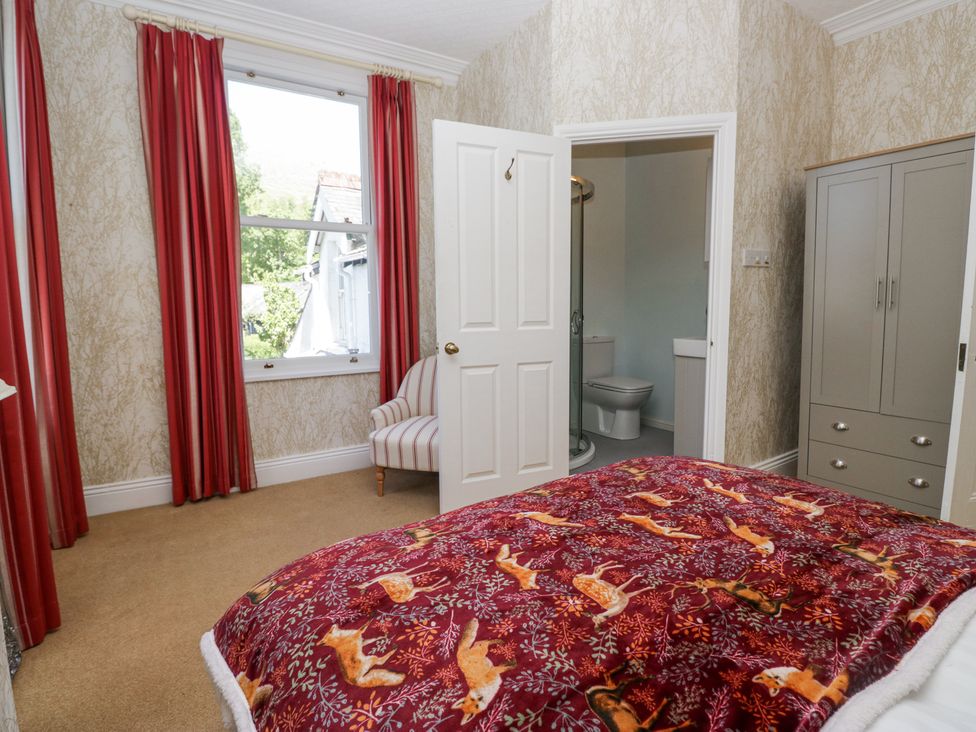 A bedroom with a bed covered by a patterned blanket a striped chair near a window with red curtains and a wardrobe next to an open door leading to a bathroom at Sunnycroft in Keswick