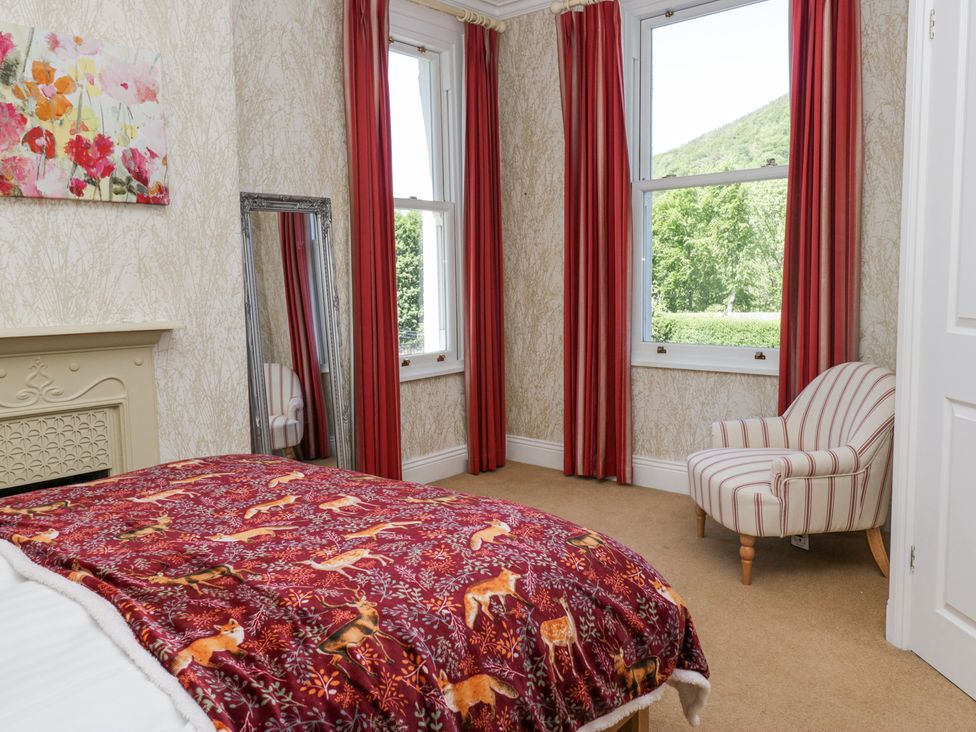 A bedroom with a bed covered in a red animal print blanket a striped chair near two windows with red curtains a tall mirror on the wall and wallpaper with a beige pattern at Sunnycroft in Keswick