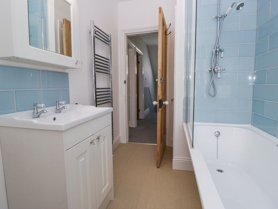 A bathroom with a white sink cabinet, a towel radiator, a wooden door, and a bathtub with blue tiled walls at Sunnycroft in Keswick