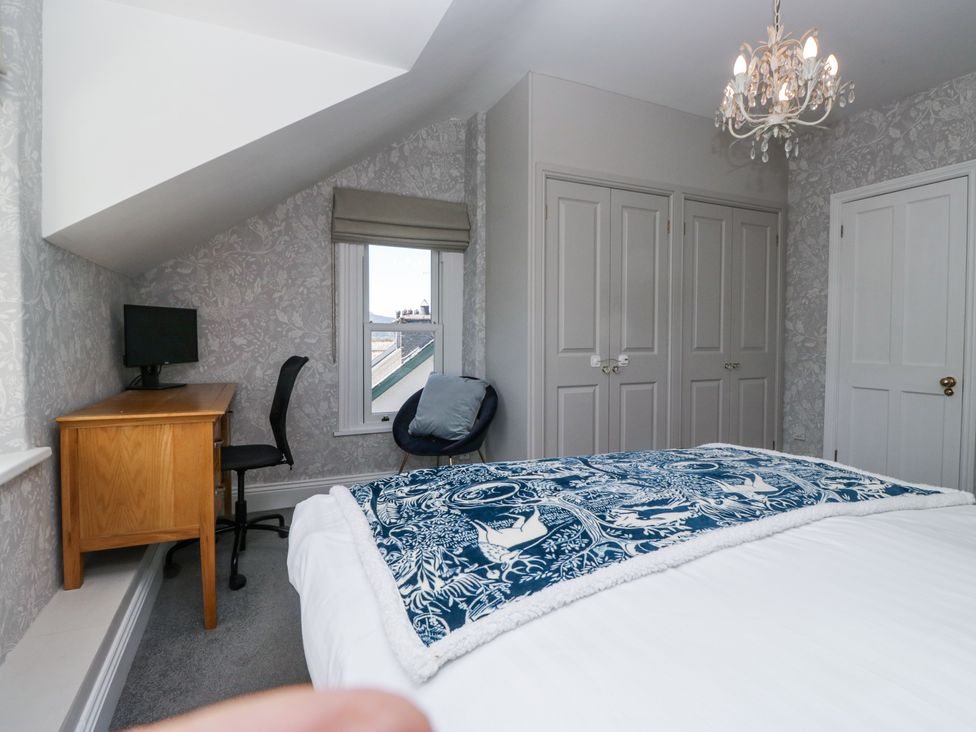 A bedroom with a bed covered with a blue patterned blanket a wooden desk with a monitor and black chair a window and built-in wardrobes at Sunnycroft in Keswick