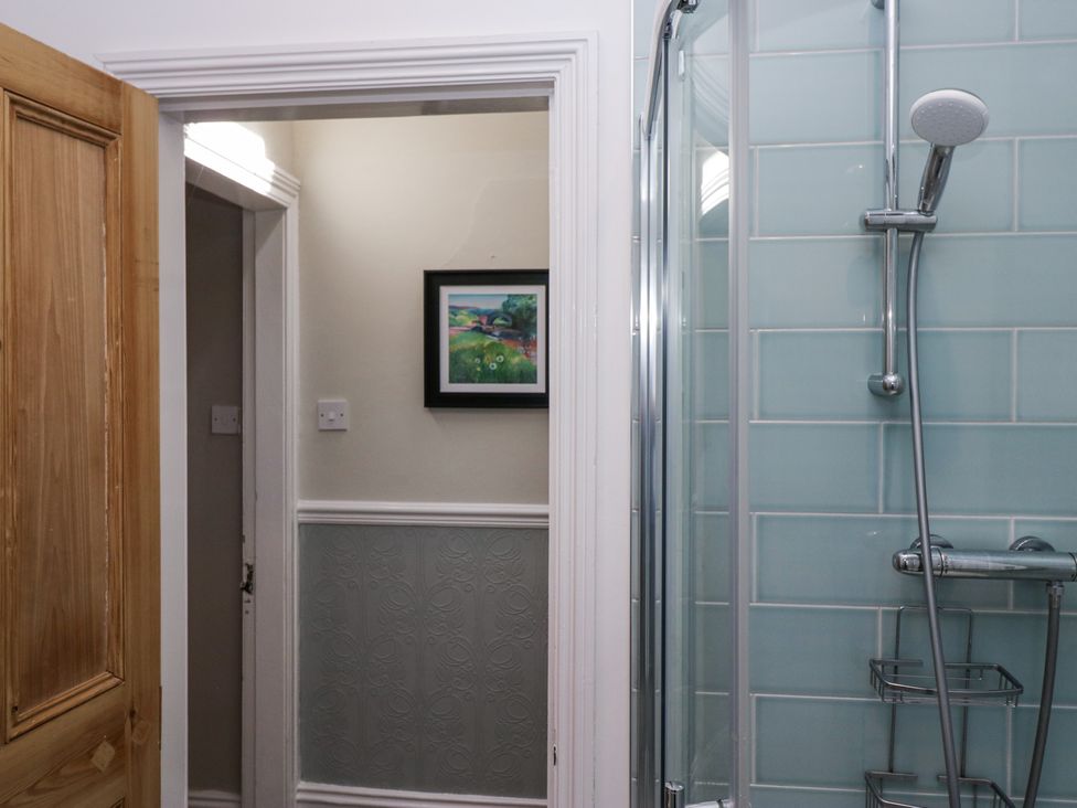 A bathroom with a glass shower enclosure with blue tiles and a wooden door leading to a hallway with a framed picture on the wall at Sunnycroft in Keswick