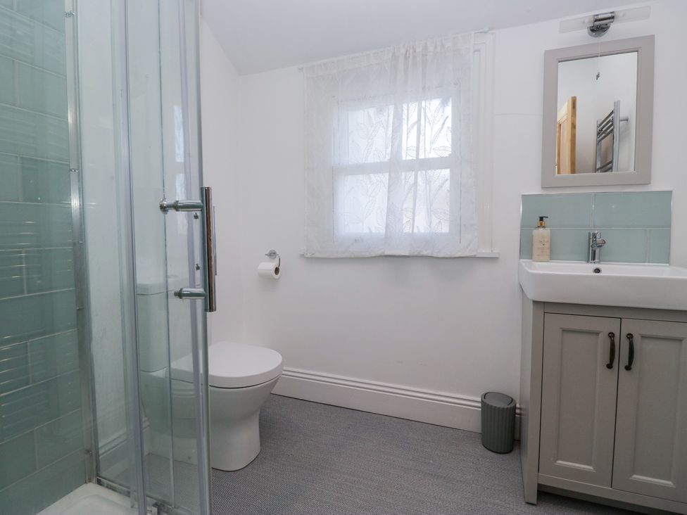A bathroom with a shower, toilet, window with curtains, sink with cabinet, mirror, and soap dispenser at Sunnycroft in Keswick