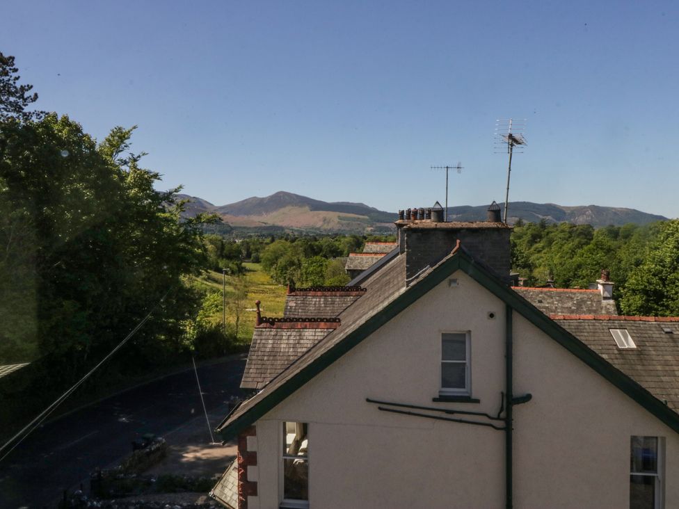 The side of a house with rooftops and chimneys visible and a road lined with trees in the background with mountains under a clear sky at Sunnycroft in Keswick
