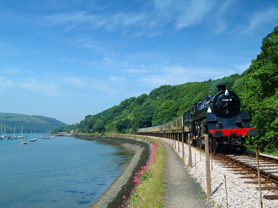 A train passing along a riverbank with boats docked in the water at 2 Court Cottage, Hillfield Village in Dartmouth