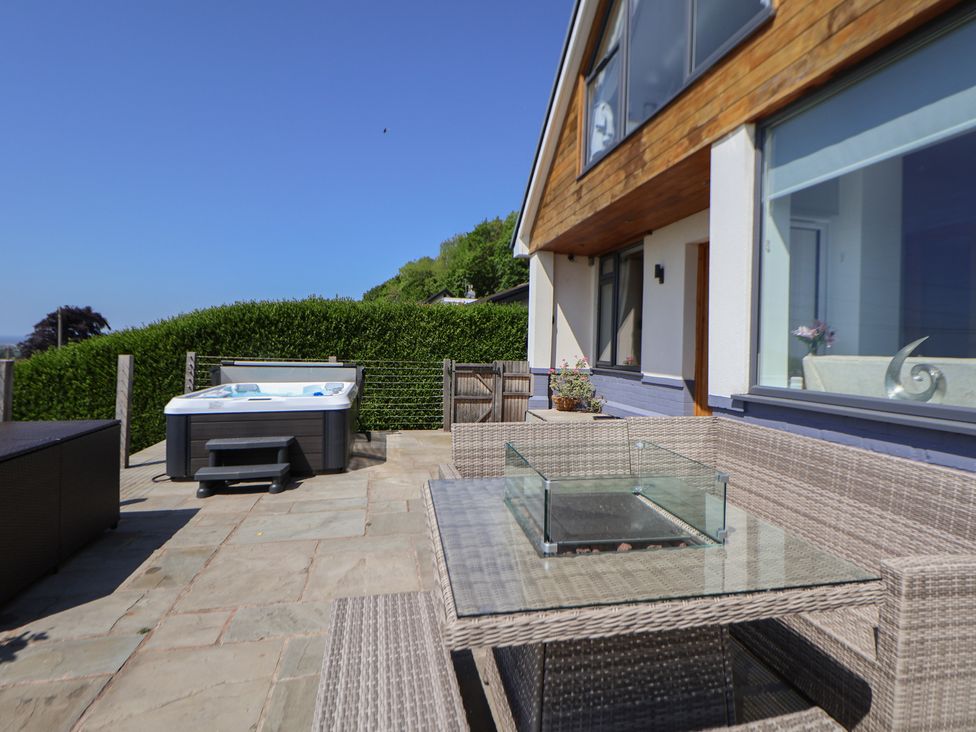 An outdoor patio with a wicker corner sofa set and glass table a hot tub and hedges at Buzzards View in Dyserth
