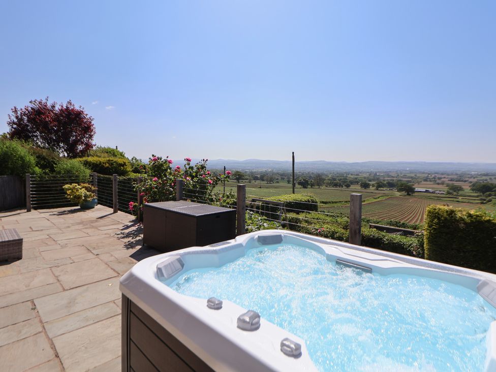 An outdoor patio with a hot tub a storage box and flower bushes overlooking fields and hills at Buzzards View in Dyserth