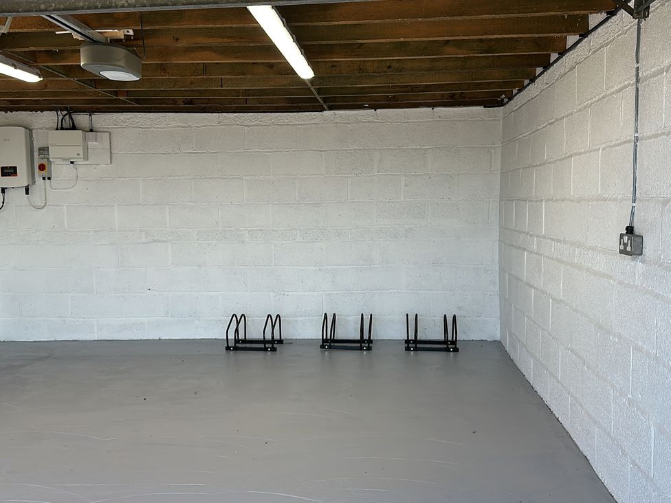 An empty room with white painted brick walls a gray floor ceiling with exposed wooden beams and three black metal bike racks attached to a wall at Buzzards View in Dyserth
