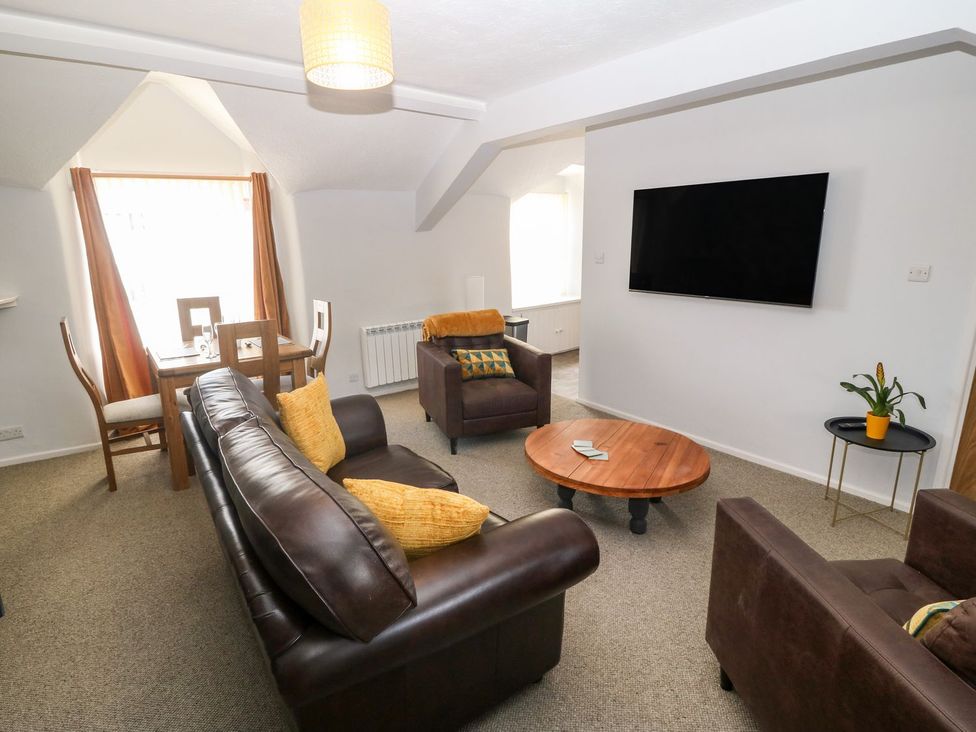 A living room with brown leather sofas a wooden coffee table a wall mounted TV a small round side table with a plant and a dining table with four chairs at Medical Hall in Cemaes Bay