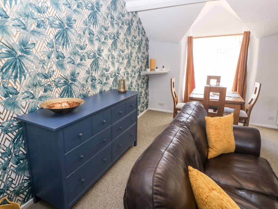 A living room with a dark leather sofa with yellow cushions a blue sideboard with decorative items and a dining table with four chairs at Medical Hall in Cemaes Bay