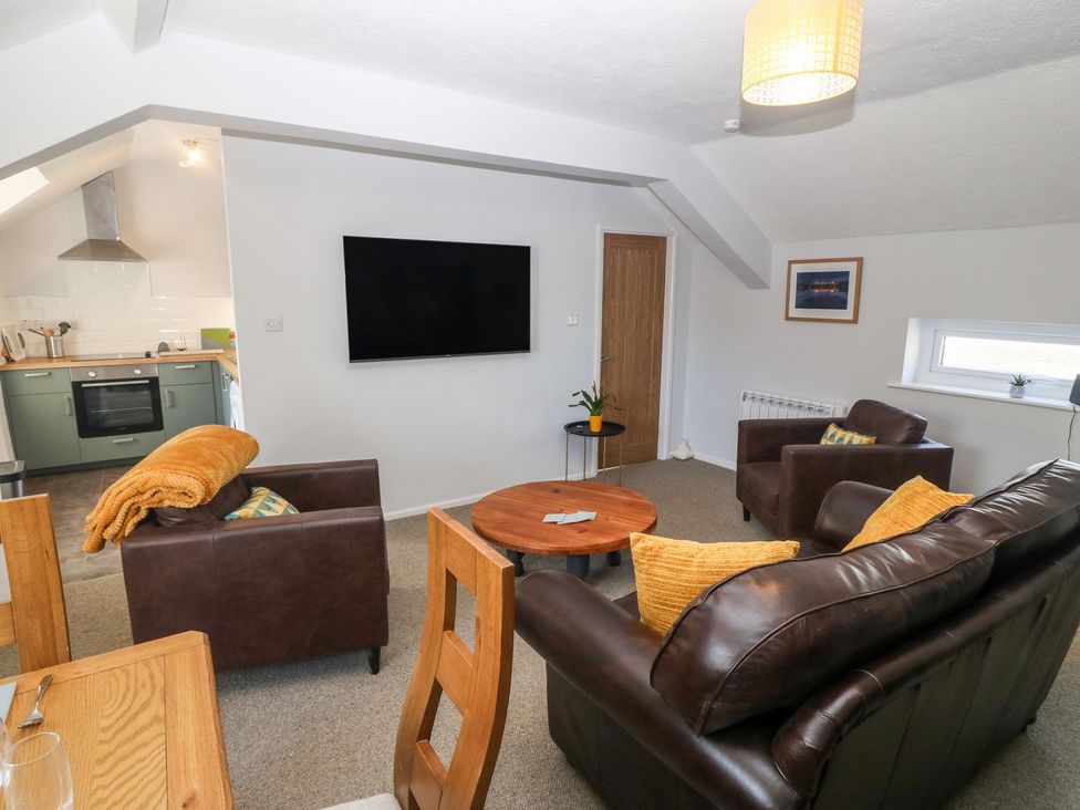 A living room with brown leather sofas orange pillows a round wooden coffee table a wall mounted television and a small dining area at Medical Hall in Cemaes Bay