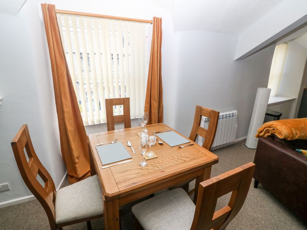 A dining area with a wooden table set for four with chairs and a window with vertical blinds and orange curtains at Medical Hall in Cemaes Bay