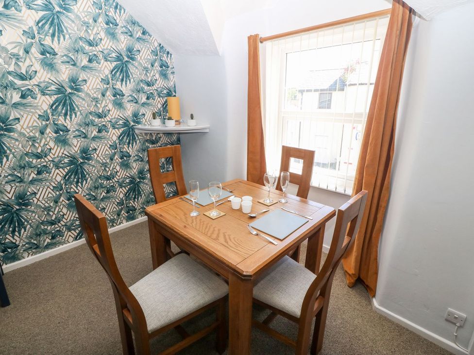 A dining room with a wooden table and four chairs near a window with orange curtains and a wall with green leaf patterned wallpaper at Medical Hall in Cemaes Bay