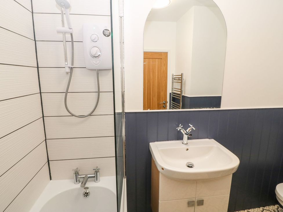 A bathroom with a bathtub and shower, a sink with cabinet below, a mirror, and a wooden door in the background at Medical Hall in Cemaes Bay