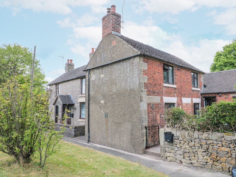 A brick and stone house with a small gated entrance and a stone wall surrounded by grass and trees at Corner Cottage in Ashbourne