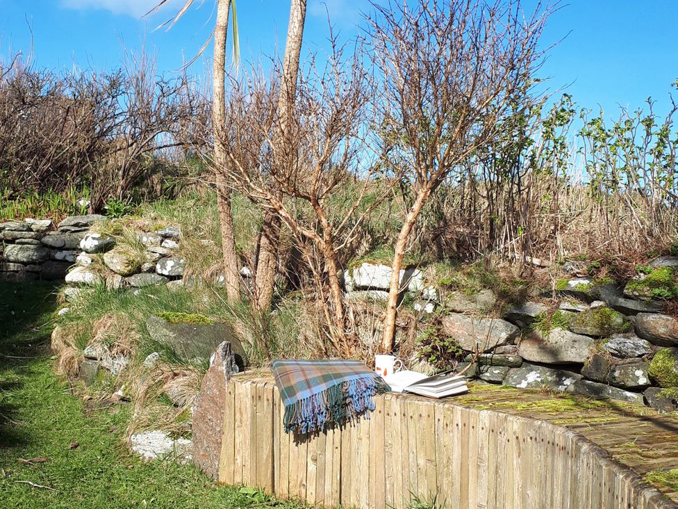 A wooden bench with a blanket, book, and mug near a stone wall and trees at Taigh Phinc, Balemore near Lochmaddy, North Uist