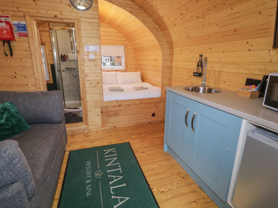 A wooden cabin interior with a gray sofa a bed shower area a blue kitchenette and a green rug at Kintala Resort and Spa