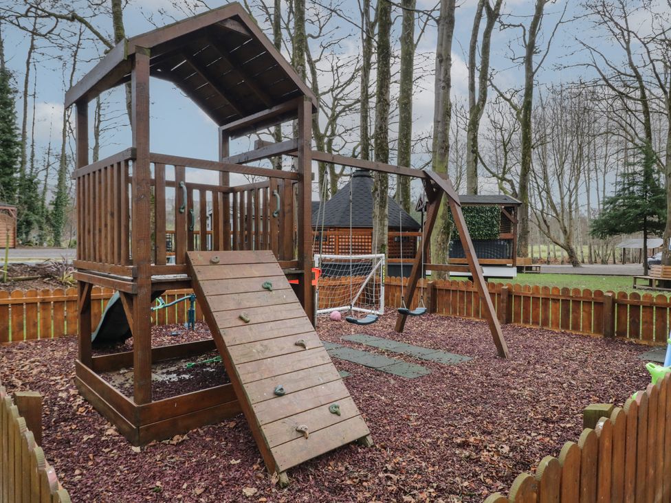 A wooden playground structure with a climbing wall swing set and slide in a fenced outdoor area surrounded by trees at Hawthorn in Dungiven Londonderry