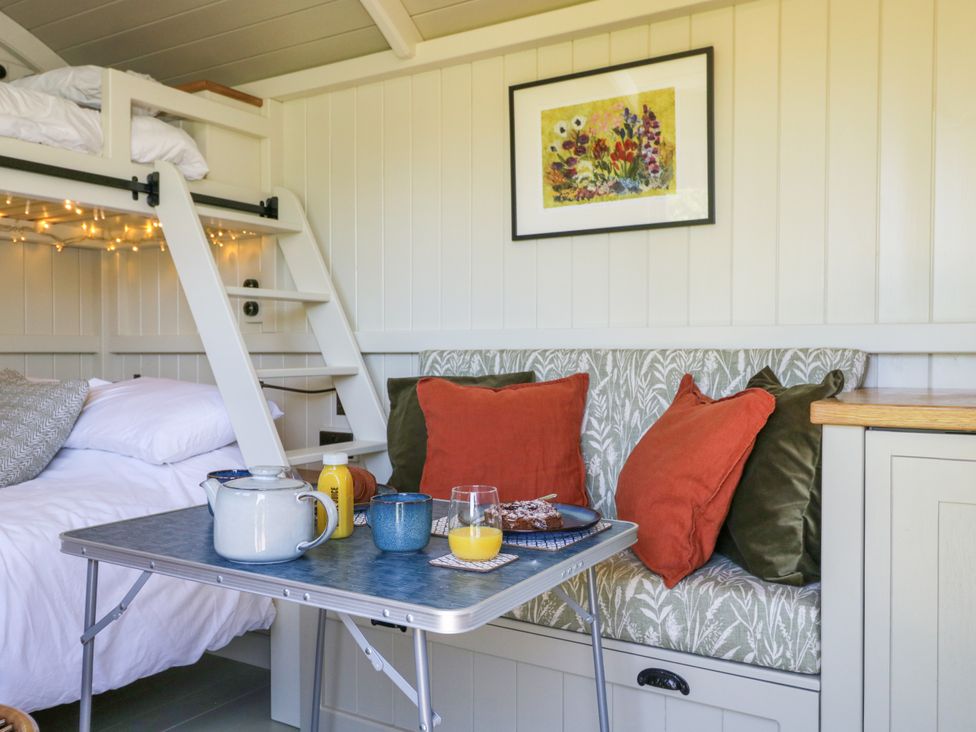 A small dining area with a table set with a teapot, cups, orange juice, and plate of food next to a cushioned bench with pillows and a bunk bed in the background at Blackthorn in South Leigh near Witney