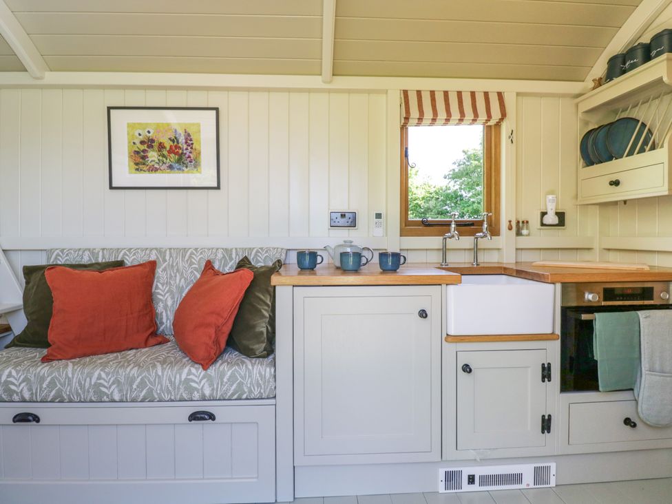 A kitchen with a built-in bench with cushions a window above a sink and shelves with plates at Blackthorn in South Leigh near Witney