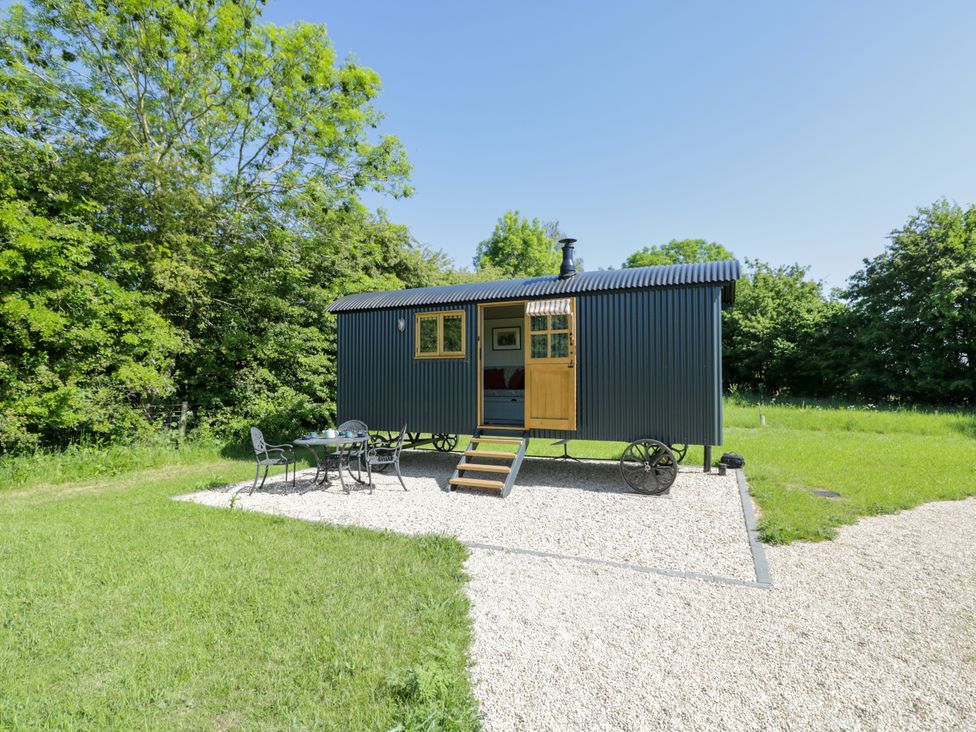 A small black corrugated structure with a wooden door and window on a gravel area with metal table and chairs surrounded by grass and trees at Blackthorn in South Leigh near Witney