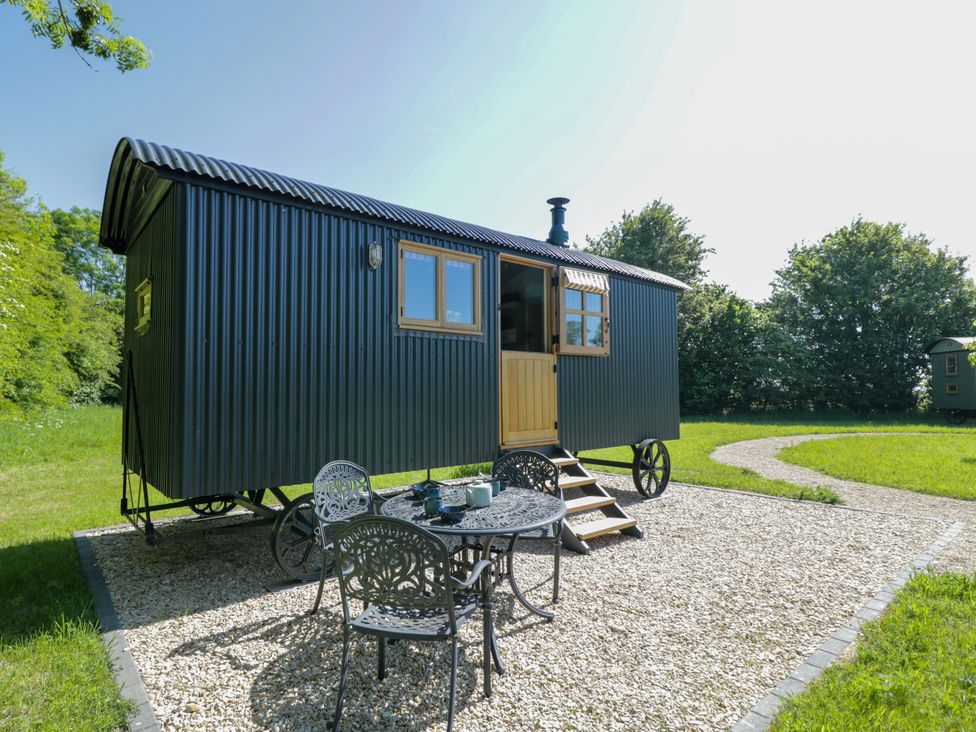 A small black corrugated metal shepherd hut with wooden door and windows on a gravel area with metal table and chairs in a green grassy outdoor setting at Blackthorn South Leigh near Witney