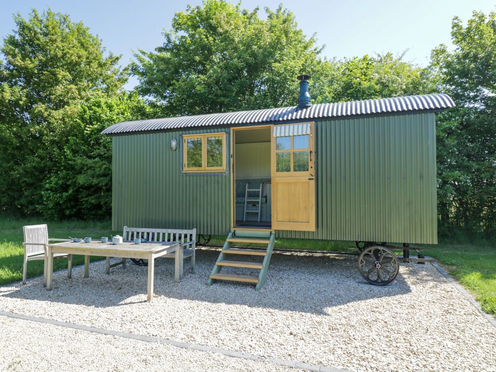 A green corrugated metal shepherd hut with open door and window on a gravel patio with wooden table chairs and bench surrounded by trees at Hazel South Leigh near Witney