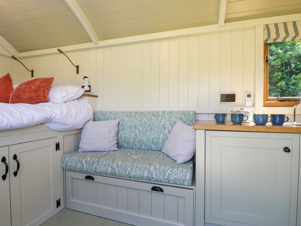 A small sitting area with a patterned upholstered bench and two cushions next to a bed with white and orange pillows and a countertop with blue mugs in Hazel South Leigh near Witney