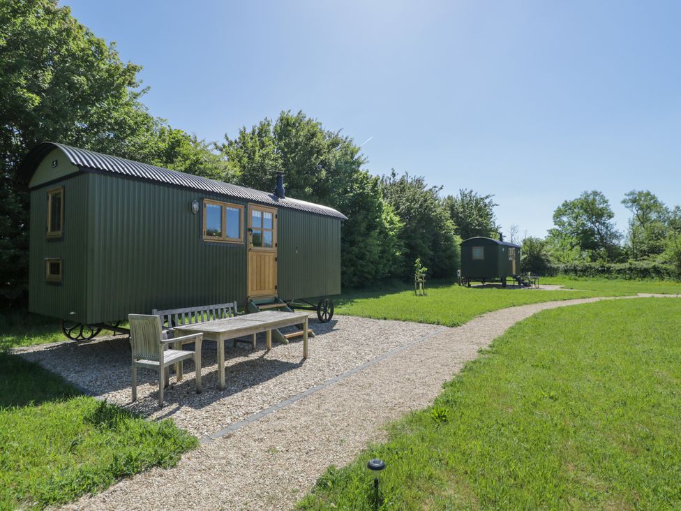 Two green metal shepherd huts with wooden doors on a grassy field with gravel paths and outdoor wooden seating at Hazel in South Leigh near Witney