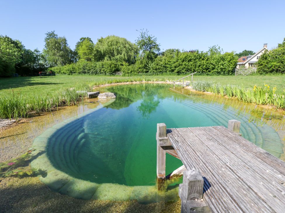 An outdoor natural swimming pond with a wooden dock in a grassy area with trees and a house in the background at Hazel South Leigh near Witney