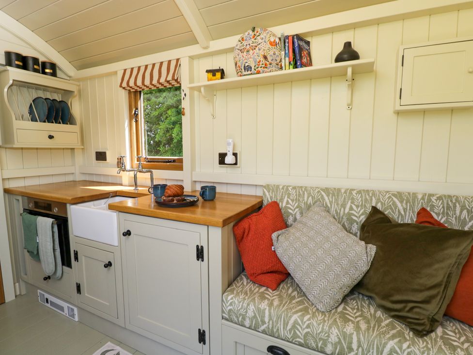 A small kitchen area with a wooden countertop a sink mugs and pastries on the counter a cushioned bench with several pillows and shelves with books and dishes