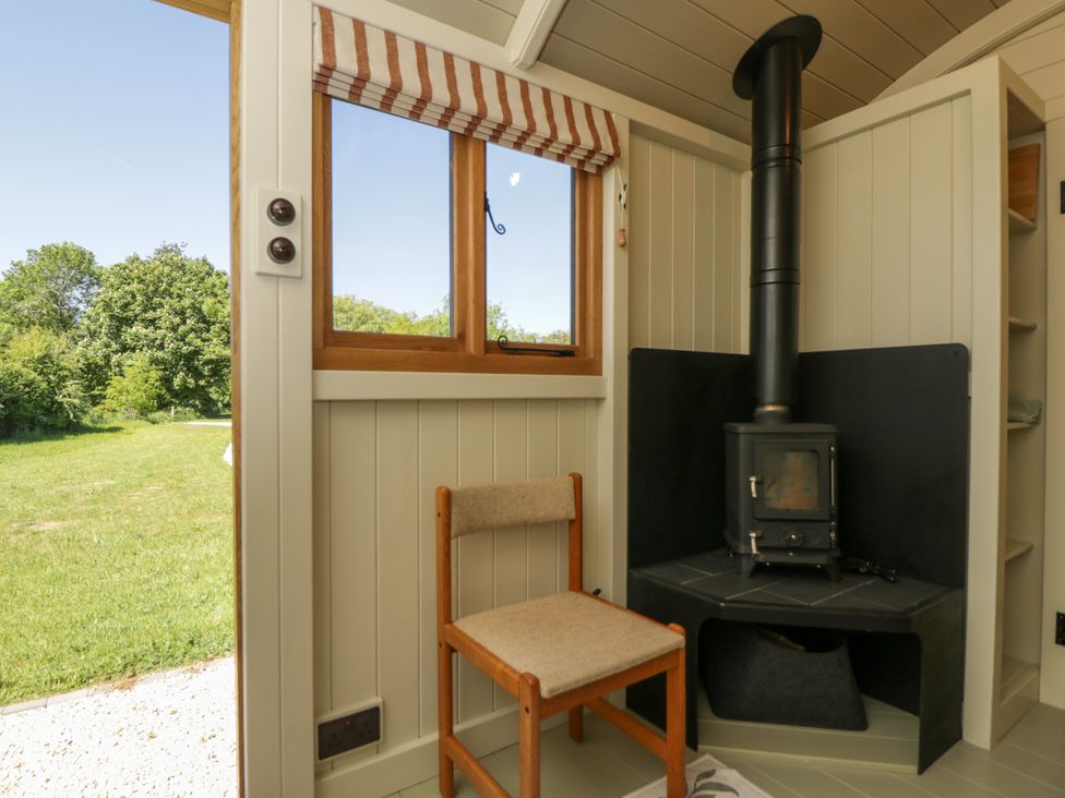 A small room with a wood burning stove in the corner and a simple wooden chair near a window overlooking a grassy yard at Spindle in South Leigh near Witney