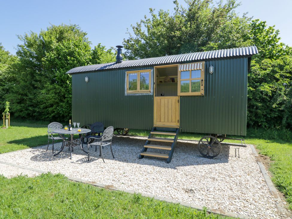 A green shepherd hut on gravel with a small black metal table and three chairs outside at Spindle South Leigh near Witney