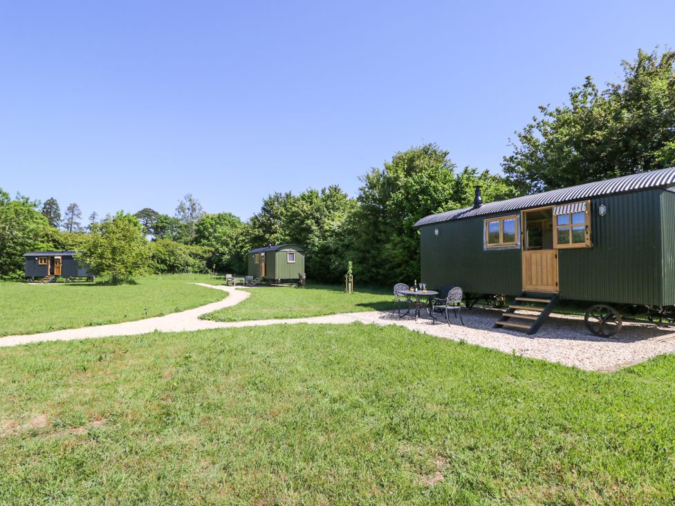 Three green shepherd huts on wheels on a grassy field with a gravel path and a table with two chairs outside one hut at Spindle South Leigh near Witney