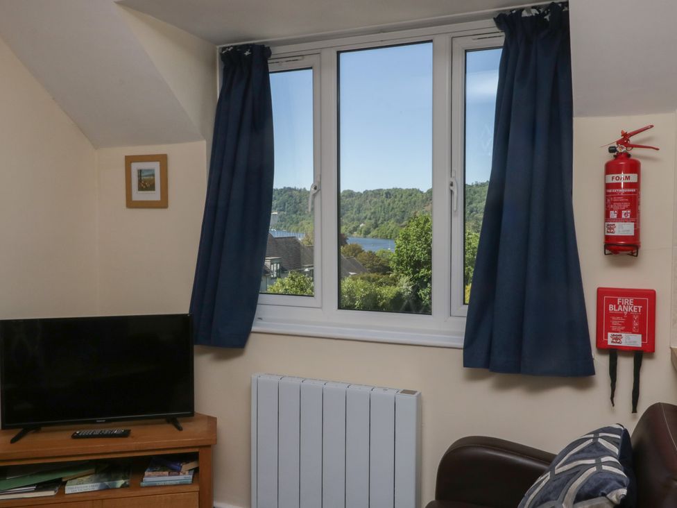 A room with a window showing a view of trees and water with blue curtains a television on a wooden stand a radiator a fire extinguisher and a fire blanket at Eve's Attic in Bowness-On-Windermere