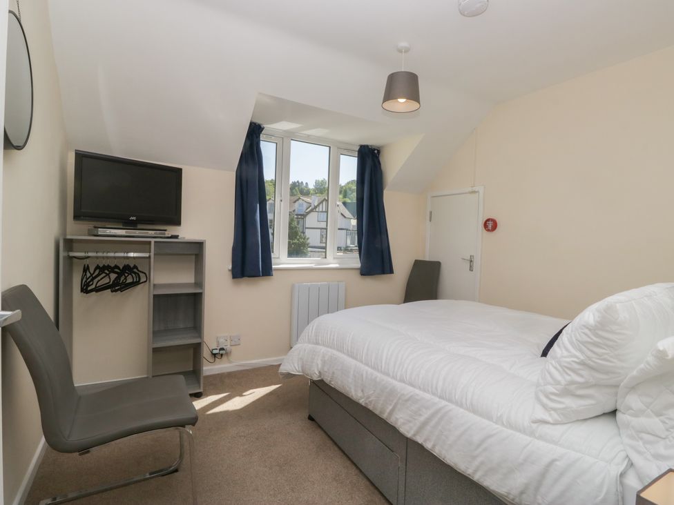 A bedroom with a double bed a chair a small shelf unit with hangers and a television near a window with blue curtains at Eve's Attic in Bowness-On-Windermere