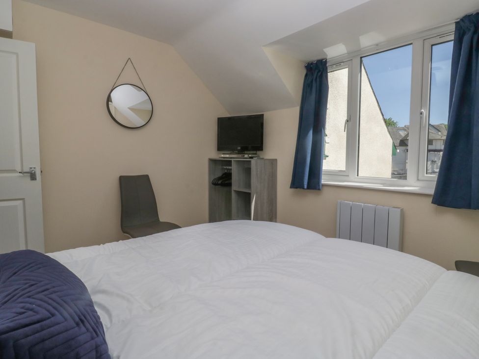 A bedroom with a bed a wall mirror a chair a small shelf with a television and window with blue curtains at Eves Attic in Bowness-On-Windermere
