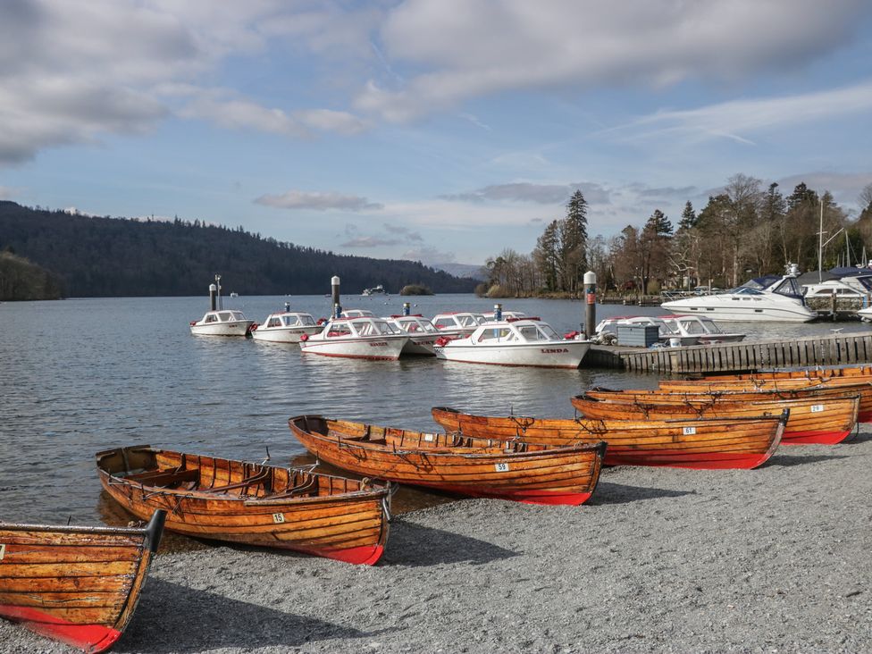 Wooden rowboats on a gravel shore next to white motorboats docked on a lake with trees and hills in the background at Eves Attic in Bowness-On-Windermere