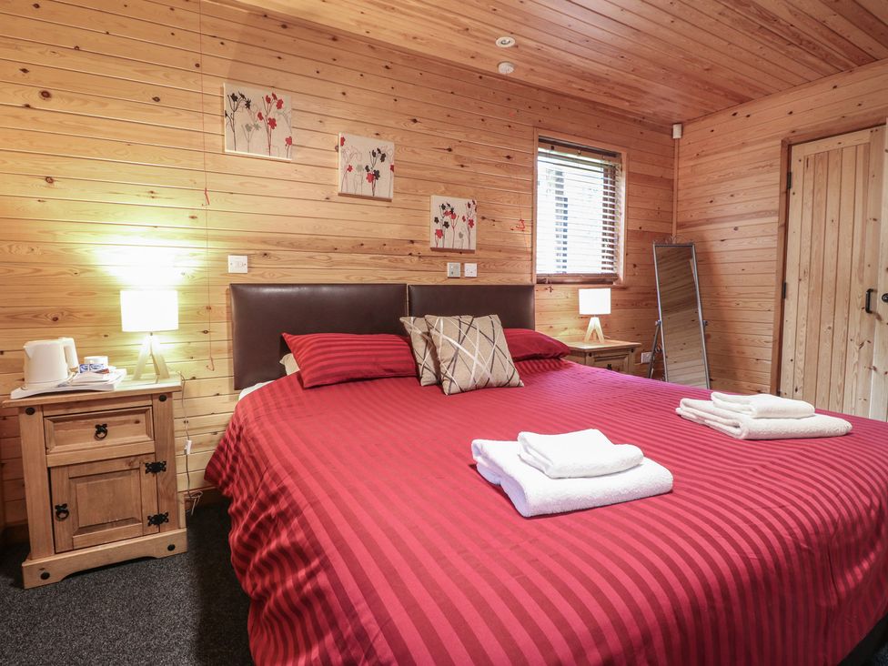 A bedroom with a wooden interior a bed with red striped bedding two bedside tables with lamps and a window with blinds at Rowan Lodge in Rosliston