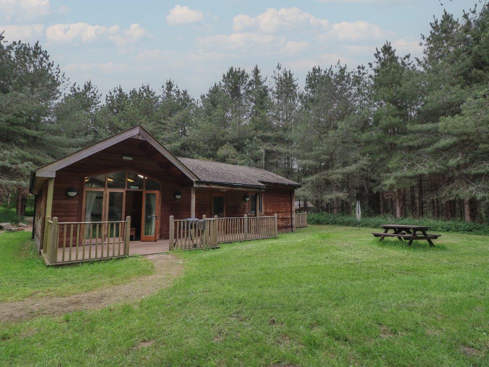 A wooden cabin with a fenced porch and picnic table on grass surrounded by trees at Rowan Lodge in Rosliston