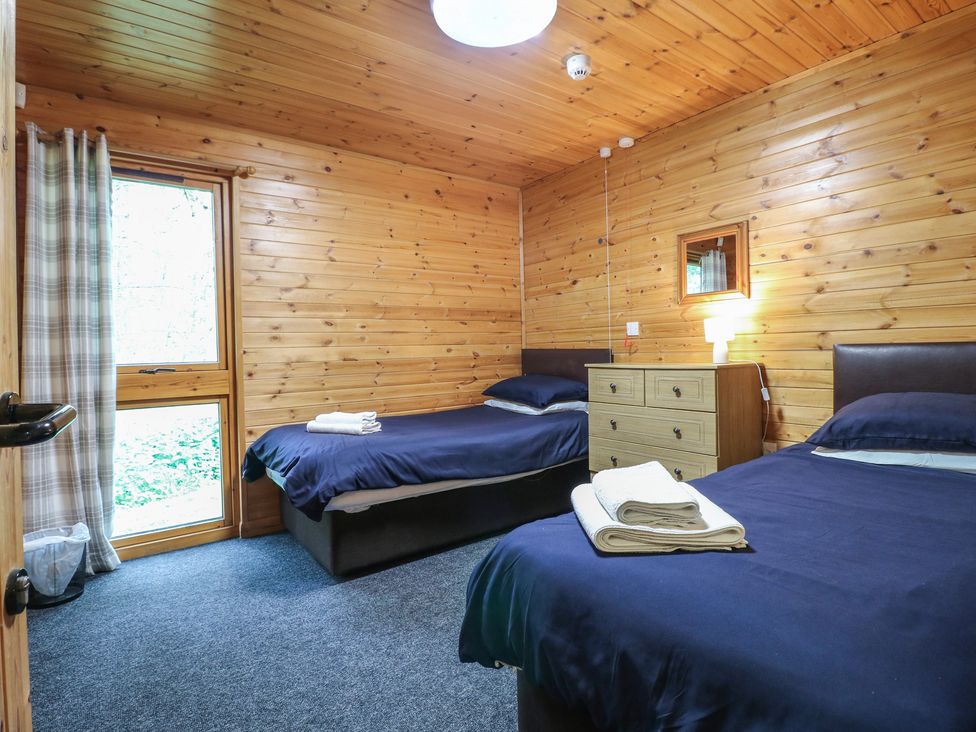 A bedroom with two single beds with blue bedding a wooden chest of drawers and a window with checked curtains at Chestnut Lodge in Rosliston