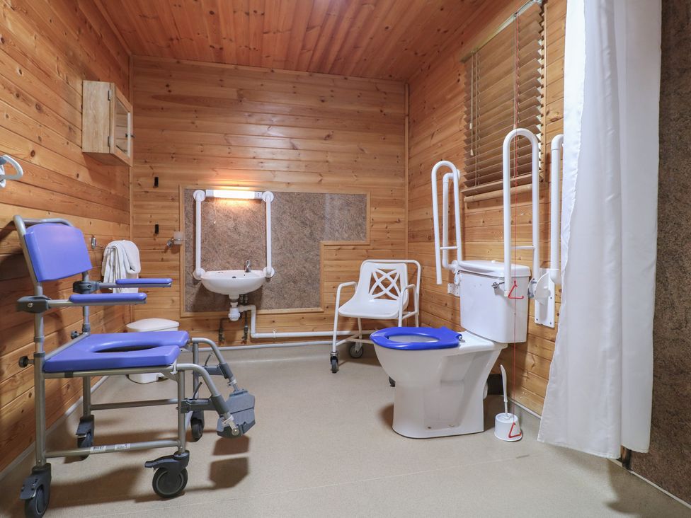 A bathroom with wooden walls a toilet with support rails a shower chair a white plastic chair and a small sink at Chestnut Lodge in Rosliston