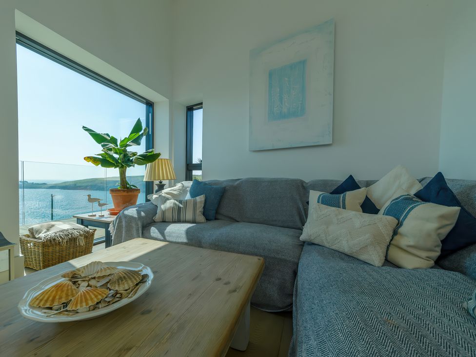 A living room with a sofa pillows coffee table and a plant by large windows overlooking the sea at Oystercatcher in Mevagissey