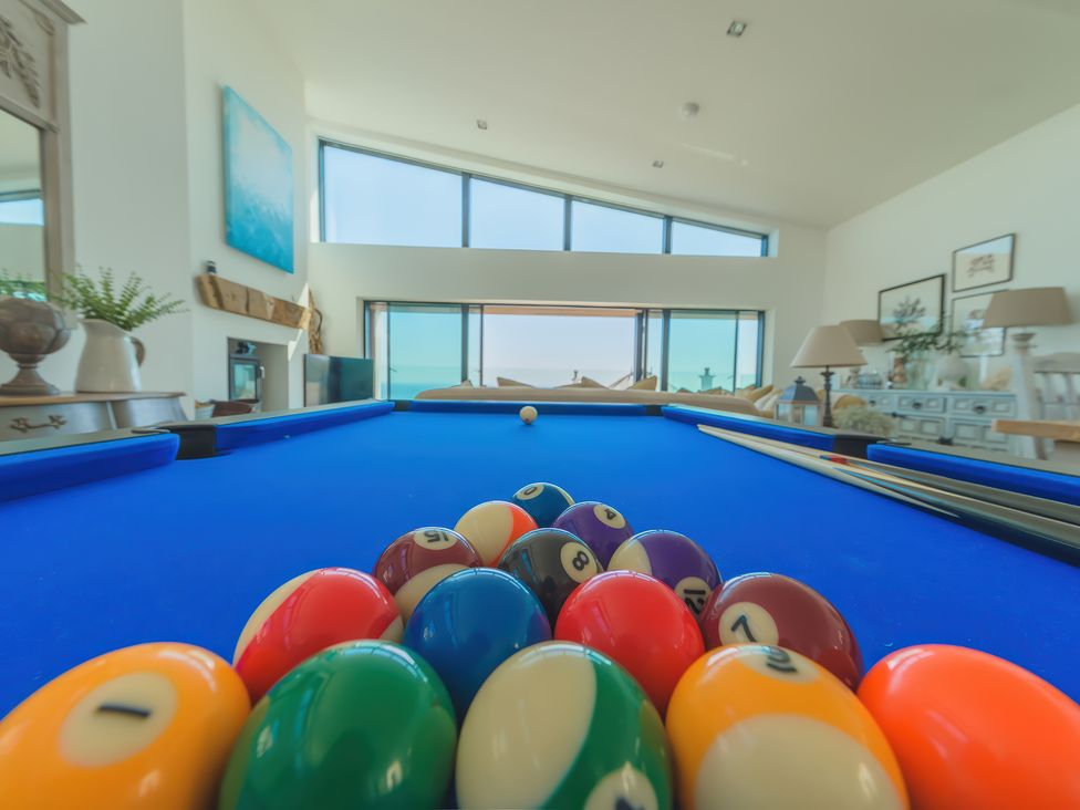 A pool table with balls racked and cues resting on the side in a room with large windows and a seating area at Oystercatcher in Mevagissey