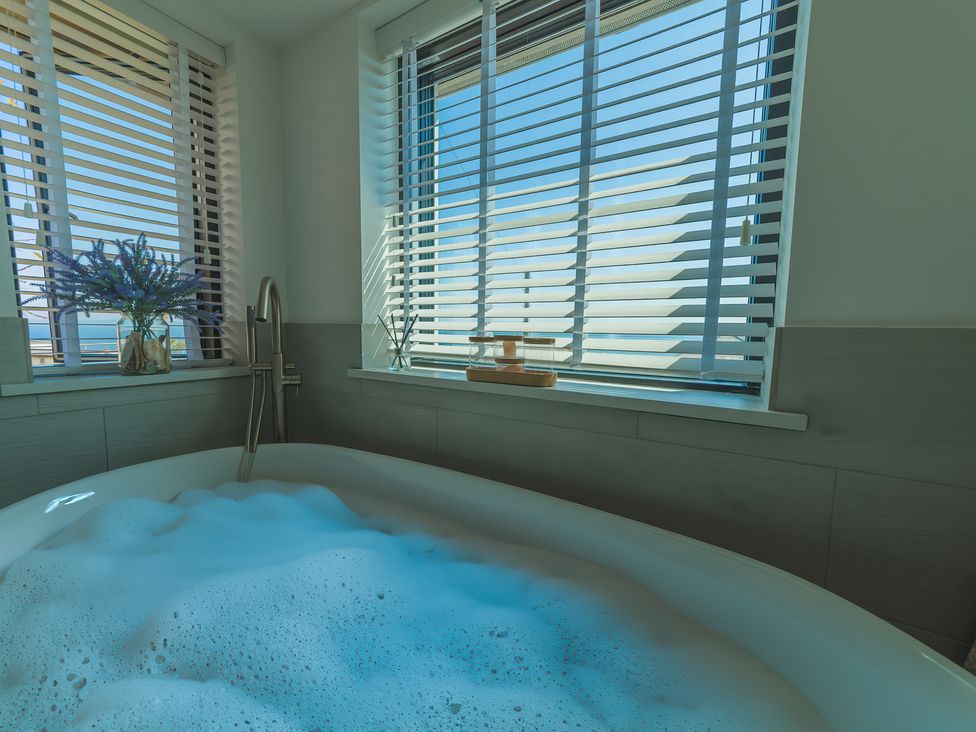A bathroom with a bathtub filled with foam and windows with blinds at Oystercatcher in Mevagissey