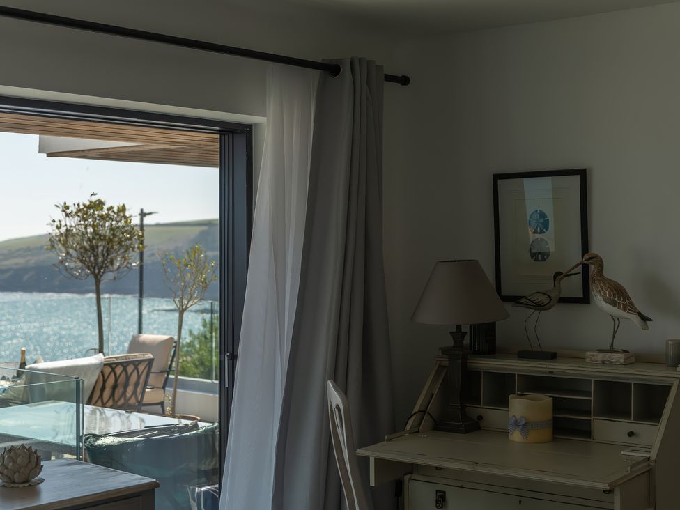 A room with a desk lamp, bird sculptures, framed picture, and a view of trees and water outside a window at Oystercatcher in Mevagissey
