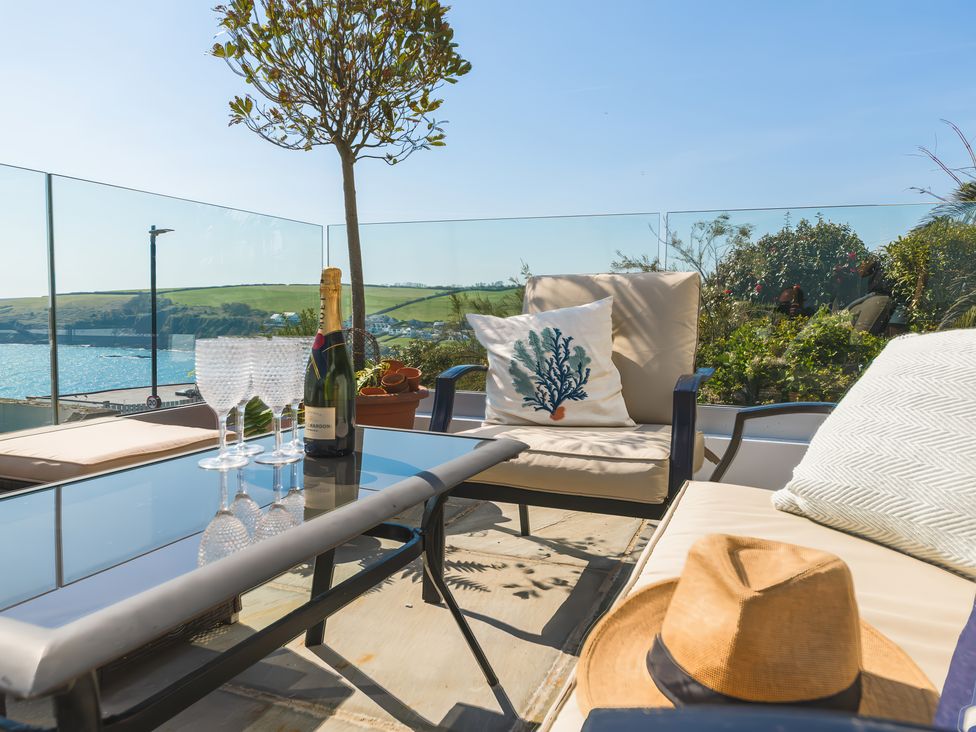 A patio with cushioned chairs a glass table with wine glasses and a bottle overlooking a coastal landscape at Oystercatcher Mevagissey