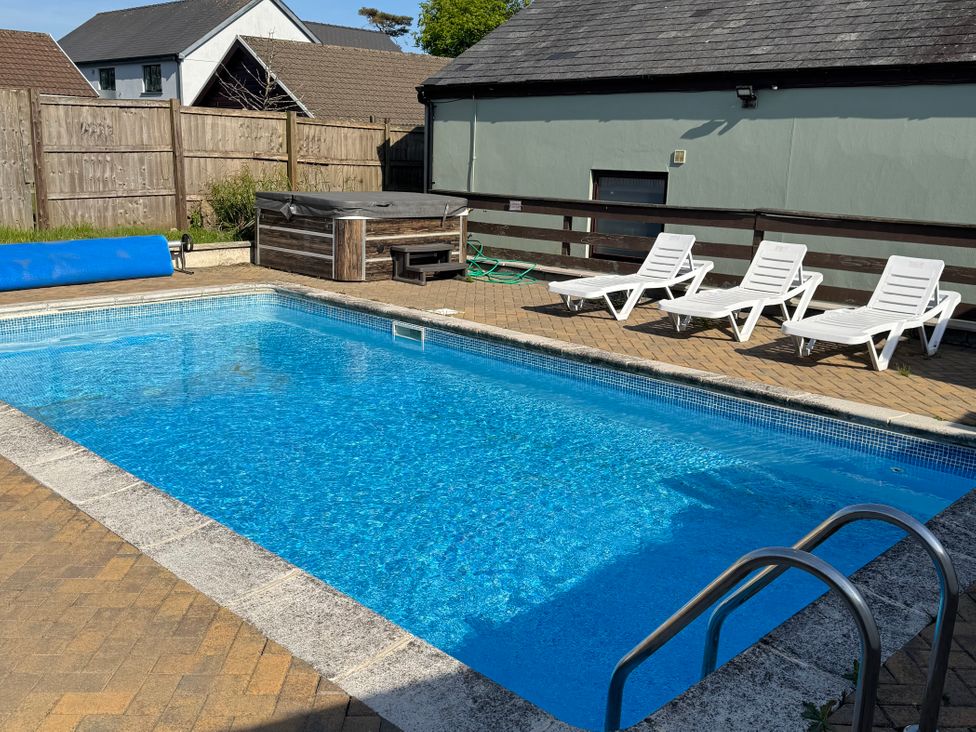 An outdoor swimming pool with a metal handrail surrounded by tiled floor and three white lounge chairs next to a wooden fence and a hot tub at Tig Bhan in Llanteg