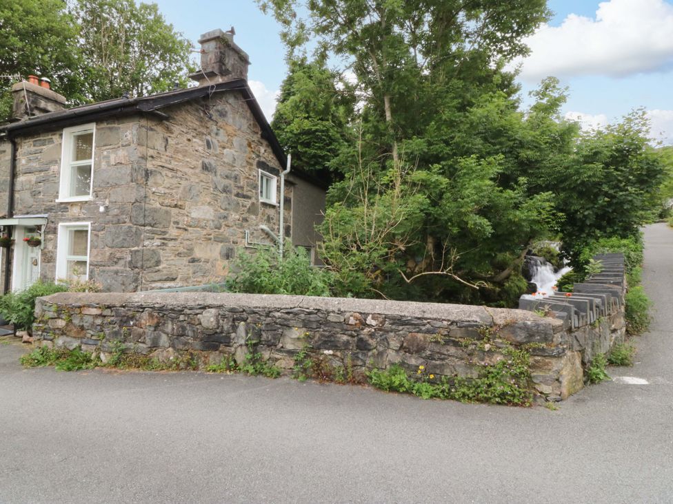 A stone house by the road with a stone wall and trees near a waterfall at Waterfall Retreat in Blaenau Ffestiniog