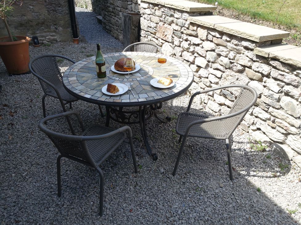 An outdoor patio with a round mosaic table set with plates of cake glasses with a drink and a bottle surrounded by wicker chairs and stone walls at 1 The Green in Silverdale